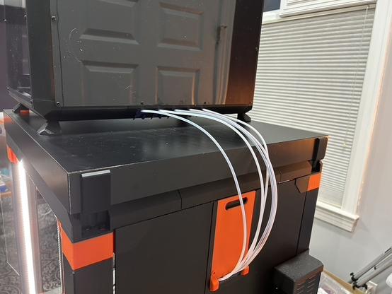The back side of the printer enclosure.  The dry box is on top.  Five white PTFE tubes emerge from the bottom edge of the box, go over the back edge of the enclosure and into an opening in the back side.  The opening is below a printed orange rectangular panel, about 100 millimeters wide by 160 millimeters tall.  At the top of the panel is an oval hole for your fingers to pull it away.  The bottom of the finger hole is black.  The panel sits flush inside a printed black surround about 12 millimeters wide.  The surround is set into a black panel that makes the rear wall of the enclosure.  The printer's power supply is mounted vertically at the very bottom of the photo.