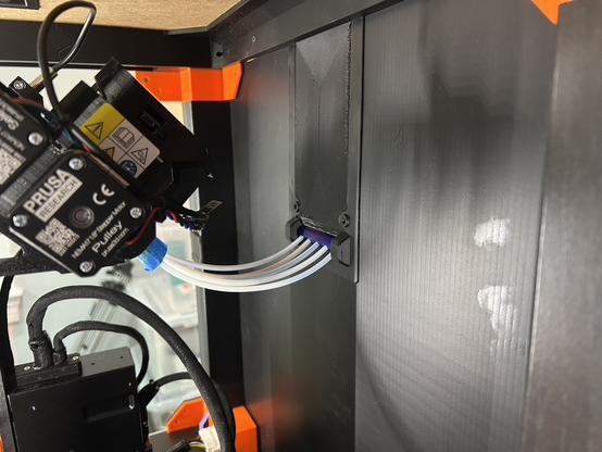 The inside of the printer enclosure at the back.  The five white PTFE tubes are coming through a purple roller, and about 12 or 14 centimeters later they are inserted into the back of the MMU.  Some of the tubes have blue tape on them.  The panel which is orange on the outside is black on this side.