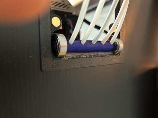 A close-up of where the tubes pass through the enclosure's back wall, viewed from outside.  The orange and black panel has been removed, and the printer is visible through the opening it covered.  The five tubes go into a purple roller.  The roller's ends are in 608 skateboard bearings, and the bearings are half-buried in the panel surround.  There are two gold disk magnets embedded int the surround about 25 millimeters above the bearings.