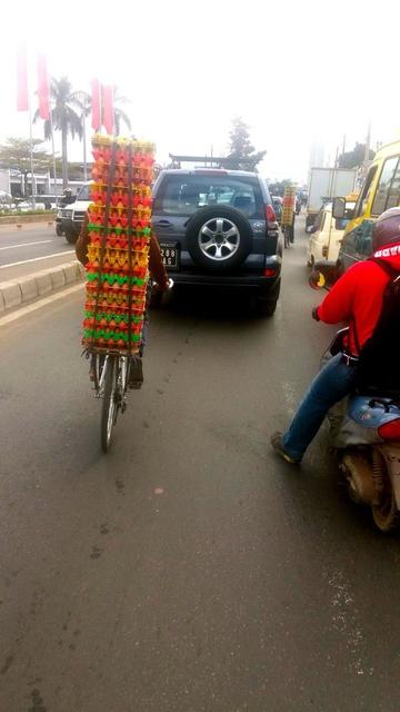 A rear view of a bicycle heavily loaded with 21 vertically stacked 6x5 egg crates, navigating through a busy traffic jam in Antananarivo.