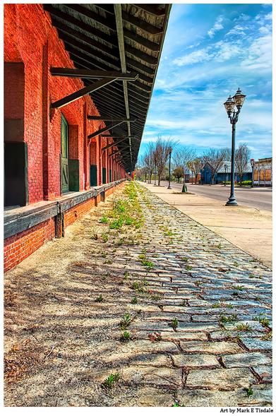 View down a sidewalk. Shadows in the foreground from an unseen winter tree. On one side of the sidewalk a quiet street. On the other an old red brick train depot with repeating arched doors for unloading freight.