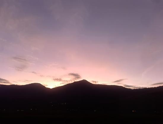 A tranquil twilight view featuring the silhouette of a rounded mountain ridge under a soft and pastel-hued sky. The colors range from pale lavender to blush pink, blending harmoniously to create a soothing gradient. Wispy clouds stretch across the sky, adding a gentle sense of movement. The foreground is dark, with tiny scattered lights from a valley below glimmering subtly, offering a connection between nature and human habitation. The atmosphere evokes a peaceful transition between day and night.