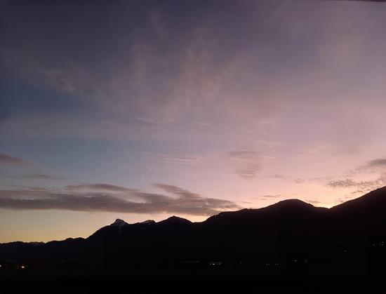 A serene evening scene capturing the silhouette of a mountain range against a dusky sky. The gradient transitions from deep purple to soft lavender and pink, with subtle wisps of clouds adding texture and depth to the composition. Hints of snow are visible on the highest peak, while faint pinpoints of light from distant structures dot the lower edges of the image, hinting at life amidst the stillness. The scene conveys tranquility and the quiet beauty of twilight.