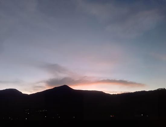 A dusky, serene sky with hues of pink and orange from the setting sun sits behind a dark silhouette of a mountain range. A few scattered lights are visible in the valley below.