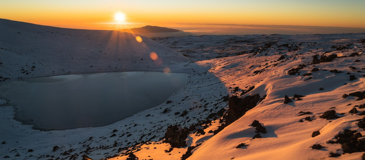 A yellow sun rising over a snowy mountainous landscape with a frigid little lake in the foreground.