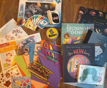 Auf dem Boden liegen im Bereich unten links verschiedene Bastelmaterialien: Aufkleber, buntes Papier, buntes Klebeband, Buntstifte und Scheren. Rechts im Bild sind Kinderbücher: „Die kleine Raupe Nimmersatt“, „Das Neinhorn“, „Im Dschungel wird gewählt“… Oben mittig liegt ein Puzzle und die Kartenspiele "Uno" und „Dobble“