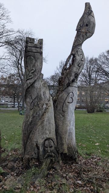 A photo of a tree trunk which splits into two. A face with a beard is carved on the bottom of the trunk. Greek letters are carved all over the tree