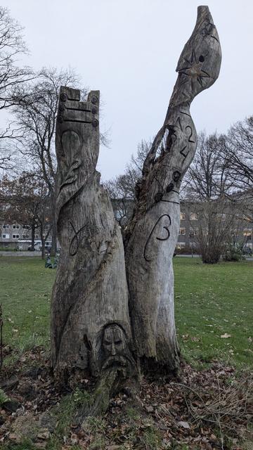 A photo of a tree trunk which splits into two. A face with a beard is carved on the bottom of the trunk. Greek letters are carved all over the tree
