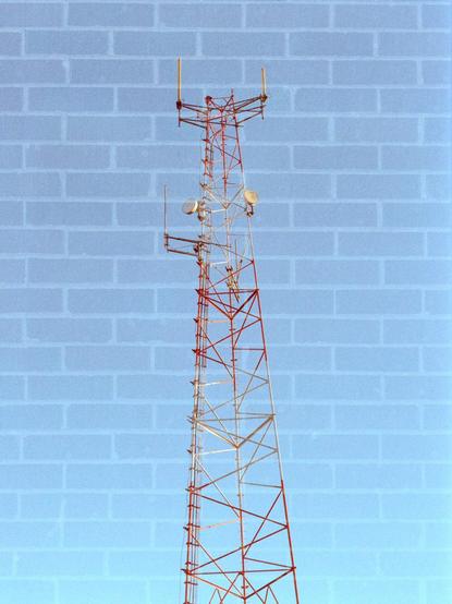 A radio tower made out of lattice construction and painted red and white with several dish and cylindrical antennas sticking out against a blue sky is super imposed on top of a brick background.