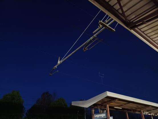 Dark blue sky with a bright white dot at the center and a few stars. Railway station platform roofs are on the upper right and lower right. A few trees are lined on the lower left.

Cielo blu scuro con un punto bianco luminoso al centro e alcune stelle. I tetti dei binari di una stazione ferroviaria si trovano in alto a destra e in basso a destra. In basso a sinistra sono allineati alcuni alberi.