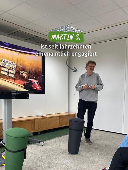 Das Bild zeigt Martin S., einen Mann mittleren Alters, der lächelnd vor einer Gruppe spricht. Er trägt ein graues Sweatshirt, dunkle Jeans und Turnschuhe. Im Raum sind moderne Möbel zu sehen: eine Holzkommode im Hintergrund, ein schwarzer und ein grüner Hocker im Vordergrund, und ein großer Bildschirm auf einem Standfuß. Der Bildschirm zeigt ein Bild von einem roten VW Käfer vor einer Mauer mit Graffiti. Über Martins Kopf steht der Text „MARTIN S. ist seit Jahrzehnten ehrenamtlich engagiert“ in einer Kombination aus grüner und weißer Schrift. Der Raum hat eine helle, freundliche Atmosphäre, die auf ein Büro oder einen Konferenzraum schließen lässt.