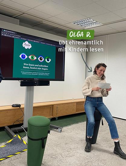 Eine junge Frau namens Olga D. sitzt lächelnd auf einem schwarzen Hocker in einem modernen Raum. Sie hält ein Blatt Papier in der Hand und trägt einen cremefarbenen Rollkragenpullover, blaue Jeans und schwarze Stiefeletten. Im Hintergrund ist ein großer Bildschirm zu sehen, auf dem eine Website mit dem Titel 'Mentor' angezeigt wird, begleitet von farbigen Augen-Illustrationen und dem Text: 'Wer lesen und schreiben kann, besitzt vier Augen.' Neben ihr ist ein grüner, zylinderförmiger Hocker zu erkennen. Olga D. engagiert sich ehrenamtlich, um mit Kindern das Lesen zu üben, wie der überlagerte Text im Bild beschreibt.