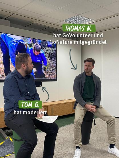 Das Bild zeigt eine Szene während einer Veranstaltung, bei der Thomas N., Mitgründer von GoVolunteer und GoNature, über seine Organisationen spricht. Er sitzt rechts auf einem schwarzen Hocker und trägt eine grüne Jacke. Links daneben moderiert Tom G., der in einem dunklen Hemd und mit Notizen in der Hand zu sehen ist. Im Hintergrund läuft eine Präsentation mit einem Bild von Freiwilligen bei der Arbeit.