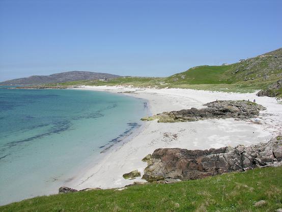 Coilleag a'Phrionnsa, or Prince's Beach, on the island of Eriskay. The image shows a white sand beach on the right and facing out onto a turquoise sea which is on the left. There is grss in the foreground and beyond the beach, on rising ground. There’s more land in the distance and rocky outcrops on the beach. The sky is blue.