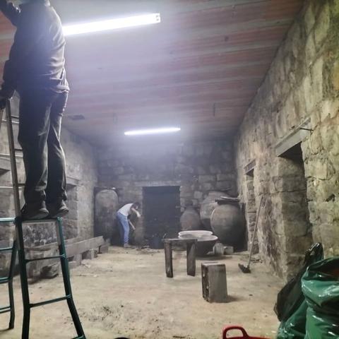 Una sala con paredes de piedra y unos fluorescentes iluminando.
Se ven unos sacos de plástico verde a la derecha. Una mesa, un cajón y varias tinajas grandes.
Hay una persona subida en una escalera en primer plano a la que no se le ve la cabeza. Y otra limpiando al fondo del todo.