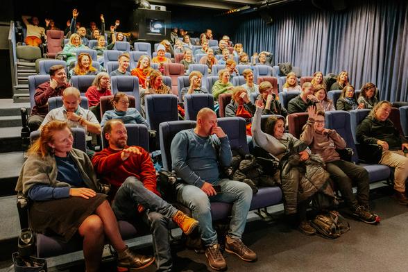 About 80 people are sitting in a cinema, the lights are on, and people are apparently having a discussion and a great time. Some of them are raising their hands.
