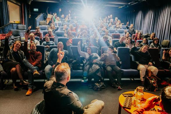 About 80 people are sitting in a cinema, the lights are on, and people are apparently having a discussion and a great time. Most of them are raising their hands. There's a contra light coming from the projector.