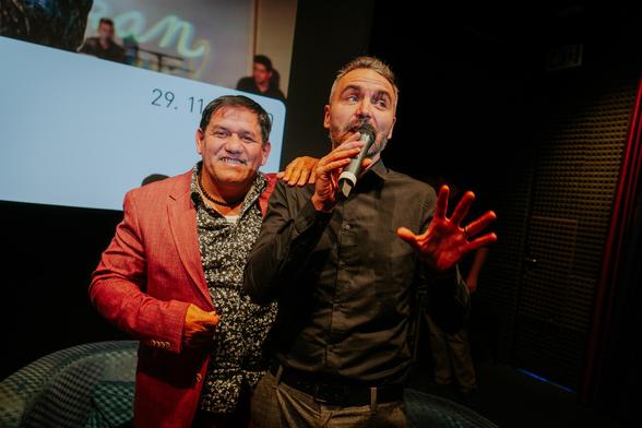 Two men in a cinema, one in a black shirt talking to a microphone, another in a red jacket smiling at the camera.