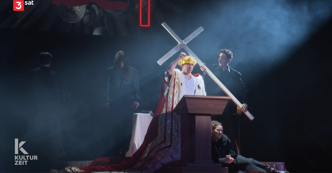 Screenshot aus der Sendung/der Inszenierung
Hinter einem Rednerpult steht ein Mann mit gelbem Haar und einem langen roten Königsmantel. Ein andere Schauspiel hält ein großes Kreuz über ihn