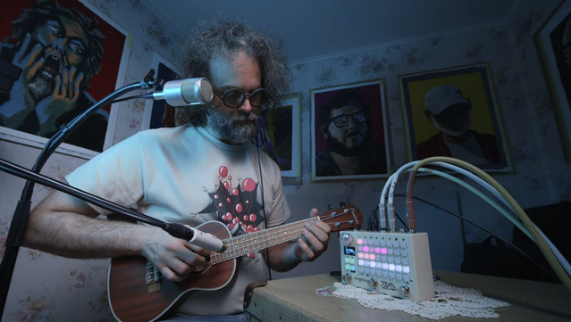 Still image of Cuckoo's video about the Zoia 10k. A White guy with chaotic black/gray hair, a full beard in the same colors, and black round rimmed glasses (Cuckoo) is sitting in a weird kind of studio set. He's playing a ukulele, pensively looking at his hands. The cable of an in-ear monitor is hanging from his ear.

In front of him is a beige kind of box, used as a table, on top of which he placed a white Empress Zoia upright on a small white crochet tablecloth. The Zoia is a small electronic device about the size of a brick, with a tiny display and a grid of RGB backlit white buttons on its front. Several chunky audio cables are plugged in at its top and go off to the side.

A mic stand, with one microphone recording the ukulele and another his voice, is standing beside Cuckoo. He's sitting in a corner in this set, which looks like an unnaturally small room with a very low ceiling. The walls have a flowery wallpaper. Five large painted portraits of people, apparently mounted on cardboard, are on the walls, and since the room is so small, they're almost touching each other. One appears to be screaming, another is looking scared, another is wearing a baseball hat and you can't see his face. Everything is tinted in cold blue light.