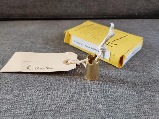 A good plated piece if lead that looks like a took chess piece. String gas been laced through it to tie a card label with a description of the artwork and the artist's name: R Claxton

Behind it is the yellow carton it came in