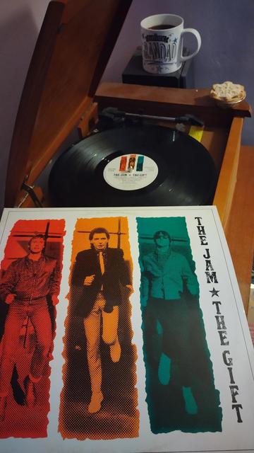 Record player with a 12" black vinyl record. In the foreground is an album cover featuring three vertical stripes, red, orange and teal, on a white background, with a member of the band in in each. Behind the player is a mug of coffee on a speaker and a delicious mince pie rests on the corner of the player.