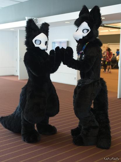 Two black creature fursuiters with white masks and blank eyes standing and clapping each others hands, in front of doors to a room where several people are congregating