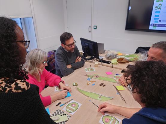 Des personnes autour d'une table travaillent sur une maquette de rivière que des castors, la mascotte Komit, doivent traverser.