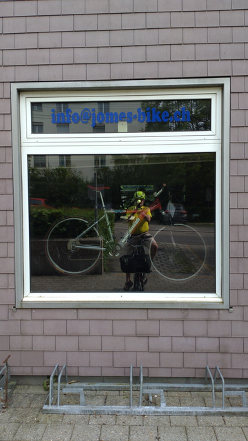 Foto durchs Fenster eines Veloladen durch. Ich spiegle mich im vorderen als auch hinteren Fenster des Ladens. Dazwischen stehen diverse Fahrräder. Ich sitze mit leuchtgelbem T-Shirt auf meinem Brompton-Faltrad.