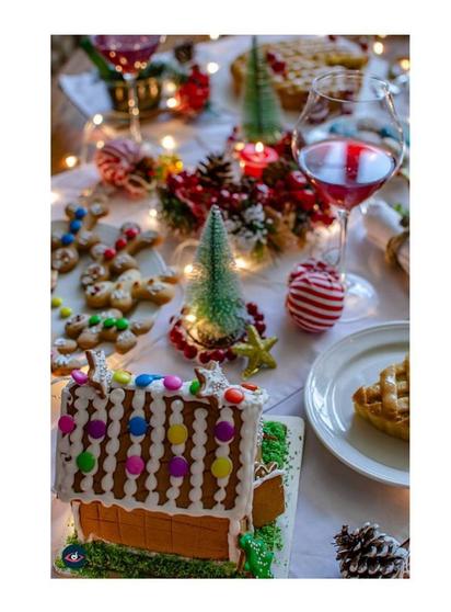 A table full of Christmas food. Gingerbread house, gingerbread men on a plate, lots of ornaments and a glass of red wine. You also see a pie.