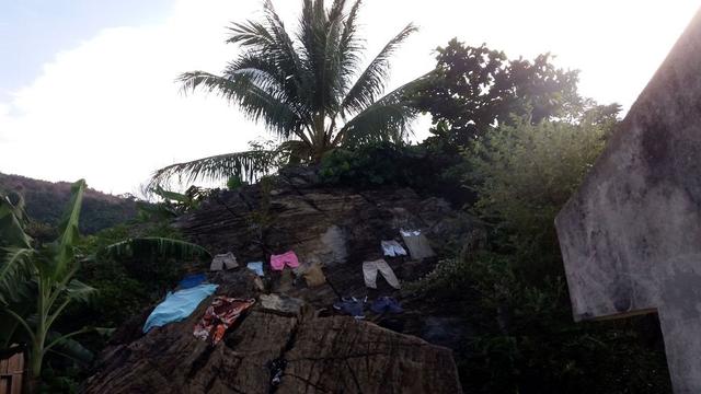 Laundry laid out on rocks to dry in Nosy Komba, with pants, shirts, and dresses arranged framed by lush greenery, including banana and palm trees.