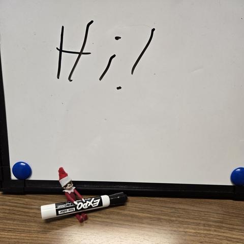 Photograph of a tiny Elf on the Shelf sitting with a dry erase marker on their lap. They sit next to a much larger dry erase board with the "Hi!" written on it very poorly.