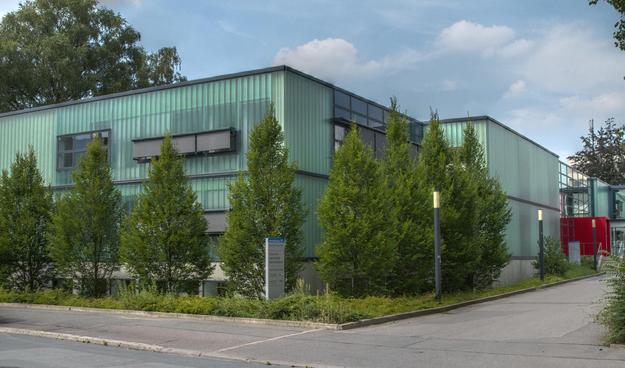 Photograph of a building under blue skies with some clouds. In front of the building are green trees. The building is covered with light-green glass bricks and has a flat roof. On the right is a bright-red entrance.