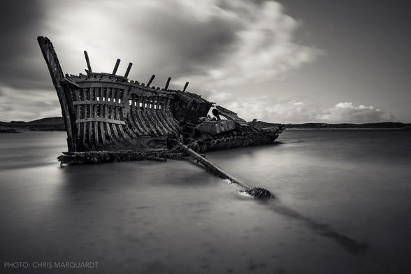 Ein altes Schiffswrack liegt im flachen Wasser. Der Himmel ist bewölkt und der Ozean ruhig, was eine melancholische Stimmung erzeugt. Das Bild ist in Schwarz-Weiß gehalten, was die Details der Szenerie betont. Interessant ist der Kontrast zwischen den scharfen Linien des Wracks und der glatten Wasseroberfläche.