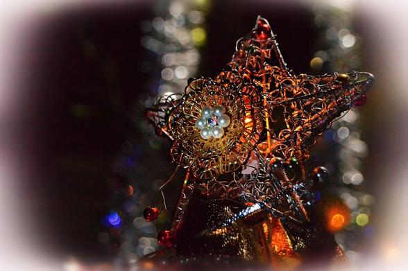 A star made of fine wire in a twisty open pattern, decorated with tiny coloured glass ornaments around the edges. In the centre a gold wire broach in a two tier leafy rosette form with a centre flower of 6 pearls around a tiny crystal. The background is mostly dark with  two silver tinsel garlands hanging down catching the light, one to each side behind the star, and with coloured lights and reflections on them. There are also a  few coloured lights and small glass balls  as well as gold ribbon below the star. A shaft of light hits the centre of the star. A filter has been applied to enhance colour intensity a bit and add a hint of violet, as well as white vignetting around the borders.