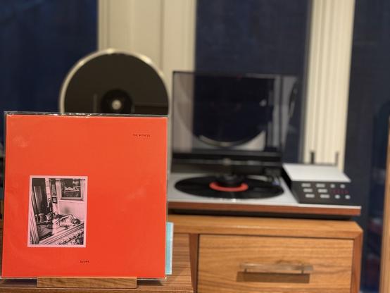 SUUNS - The WItness LP cover.

An orange/red cover with a small, inset black-and-white photo of a naked man, lying on a bed, in front of a wall that has a picture of a woman, facing away, naked on a bed.

The black LP plays on a vintage, restored Bang & Olufsen Beogram 8002 linear-tracking turntable to the right.