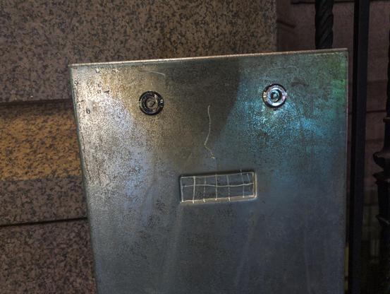 A metal utility bollard looks like a worried face due to two unlocking rings level with each other, and below them a nose drawn in white, and in a rectangular recess with teeth drawn in white.