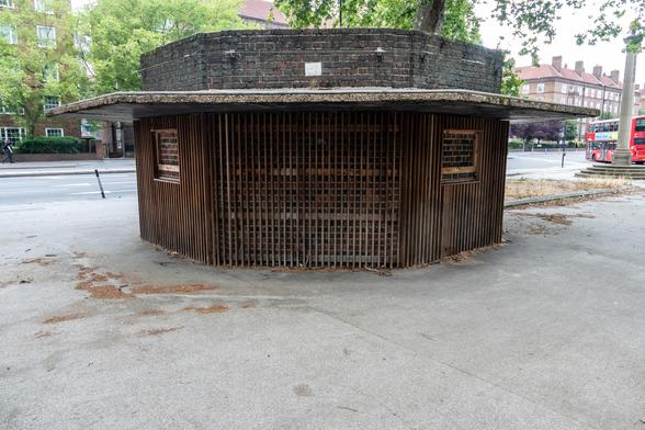 A hexagonal brick building looks like a head wearing a hat due to a wide rain shelter extending out from it, two windows that have brick behind them, and a wide grille area.