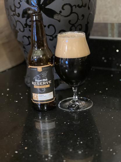 A tall tulip glass with a huge tan head filling 1/3 of the glass. The brown bottle to the left has a black label with orange edging, white letters, and a dragon with furled wings in silhouette