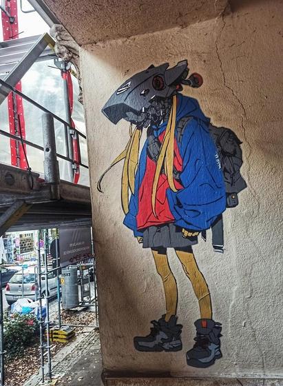 Streetartwall. A paste-up with a drawing of a robot man was stuck on a beige-colored house wall, somewhat hidden behind scaffolding with tarpaulins. The self-designed paste-up shows a futuristic vision of a human hybrid with a large camera for a head. The figure has long blond strands of hair, wears a red hooded jacket with very wide blue sleeves, short gray shorts, yellow leggings, wide gray sports shoes and a backpack. Very stylish and unusual.