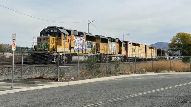 Two heavily graffitied yellow locomotives pulling a few flatcars stacked with wood cargo idles on a single track behind a fence on a cloudy day.