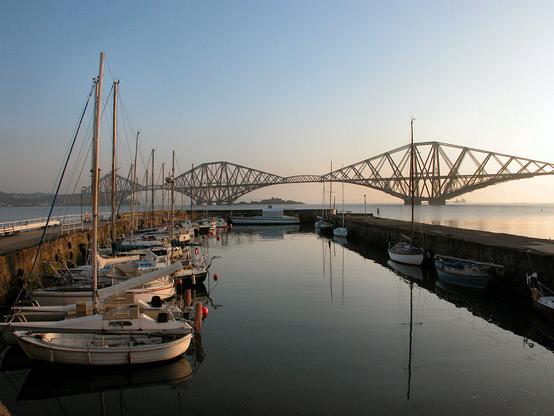 The Forth Bridge, also known as the Forth Rail Bridge, seen from the harbour at Queensferry or South Queensferry. The harbour occupies the bottom half of the frame and has walls on both sides sticking out into the Firth of Forth. The harbour is very still and reflective and home to lots of leisure boats on the left. The Forth Bridge extends across the middle of the frame. The sky is blue in early morning light.