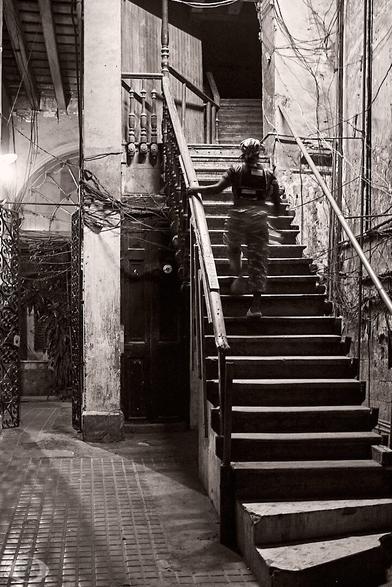 B/W photo of a woman going upstairs. The stairs occupy the right half of the photo ending up in a door with a balcony and continuing after that. Electric wires crawling around the wall and ceiling, which would make a firefighter get an immediate heart attack.