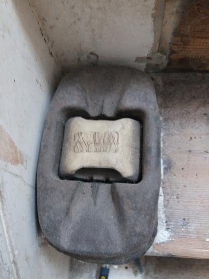 A cats eye reflector removed from a road. It is a large rectangular lump of cast iron, slightly rounded with a profiled top. In the centre is a white rubber insert that holds a pair of reflective cats eyes at each end. The profle of the cast iron allows the cats eye to be safely driven over but the 'eyes' to still be seen by approaching traffic.
It is on the end of a wooden stair case tread agist the stringer. The stair needs repainting and carpeting.