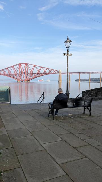 A man sitting on a bench looking at the Forth Bridge. The water is very calm and there's an almost perfect reflection of the bridge. There's a mistiness to the sky. It's hard to see where the water ends and the sky begins.