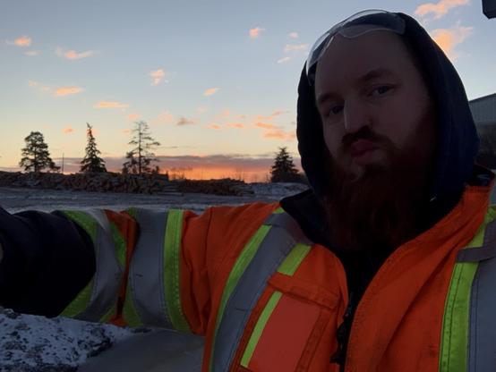 A husky cis male with a big red beard offers a virtual hug. He’s wearing a bright reflective jacket. The sun is rising in the background