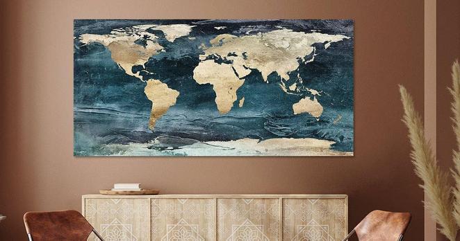 A mock-up of a decorative world map art print in ocean blue and sand with lots of texture. The mock-up is shown in a rustic room with light brown walls, a chair and a natural wood sideboard.