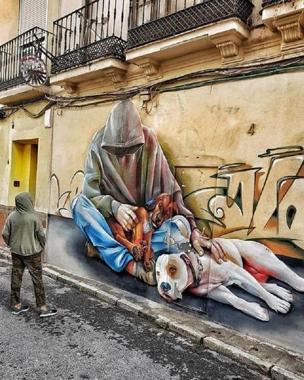 A mural by Lalone in Málaga, Spain, featuring a hooded figure sitting on the street with two dogs. One dog lies resting on the ground, while the other is curled up in the figure's arms. The mural combines photorealistic details with graffiti-style accents.