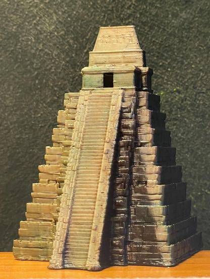 Miniatura impresa en 3d y pintada a mano del templo del Jaguar en Tikal (Guatemala). La foto lleva luz cenital por lo que las luces y sombras que se aprecian son producto de la pintura y no de la iluminación. Foto frontal; la miniatura está frente a un fondo verde oscuro, y sobre una tabla de madera. En esta foto se aprecia muy bien como las distintas estructuras proyectan su sombra sobre las demás.