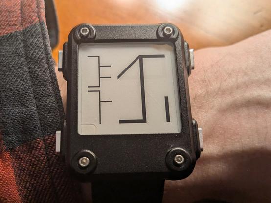 A photograph of a Watchy - an e-paper smartwatch with a black metal case - showing three Cistercian numerals on screen. A large one on the right has a value of 1631, meaning that it's 16:33 in the afternoon. There are two smaller numerals stacked vertically to the left of it. The upper one has a value of 1212, meaning that it's the 12th of December. The lower one has a value of 12, indicating that I've taken twelve steps since flashing this code to the watch.